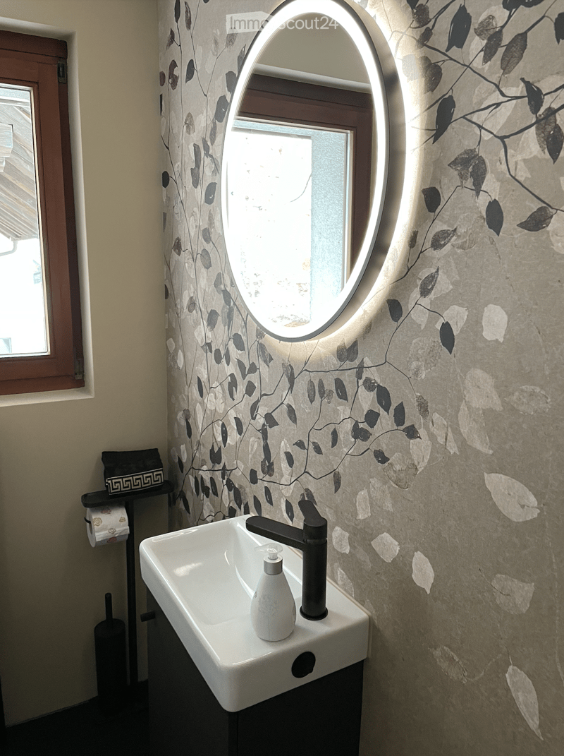 Modern bathroom with a white sink, black faucet, soap dispenser, illuminated round mirror, and decorative leaf-patterned wallpaper.