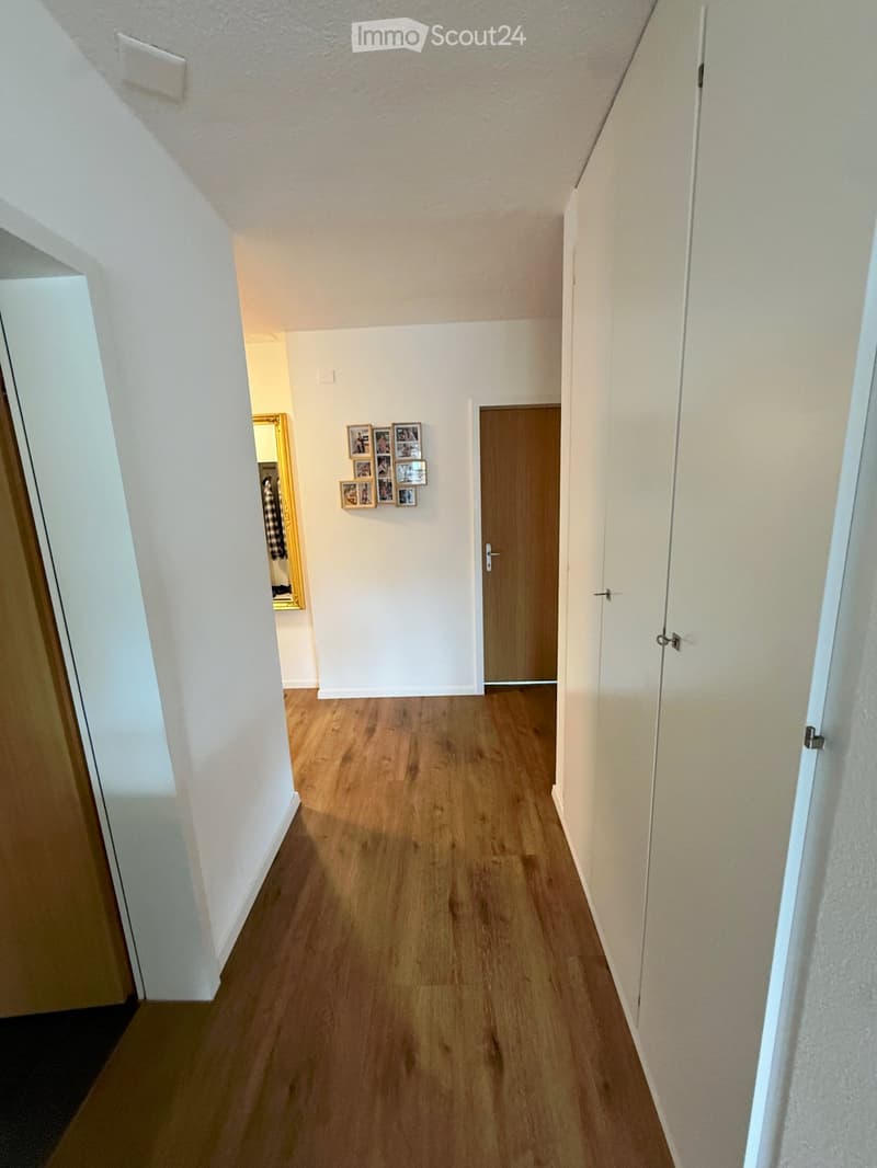 A narrow hallway with wooden flooring, white walls, and several doors leading to other rooms. A small photo collage is mounted on one wall.