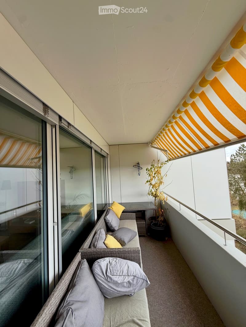 Balcony with a wicker sofa, cushions, small table, and potted plant, covered by a striped awning.