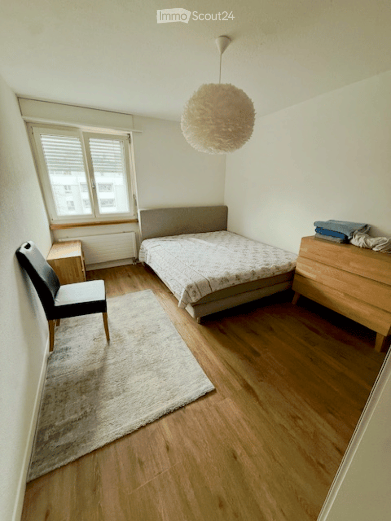 Cozy bedroom with wooden flooring, a bed with a grey headboard, a nightstand, a wooden dresser, a chair, a round rug, and a hanging pendant light.