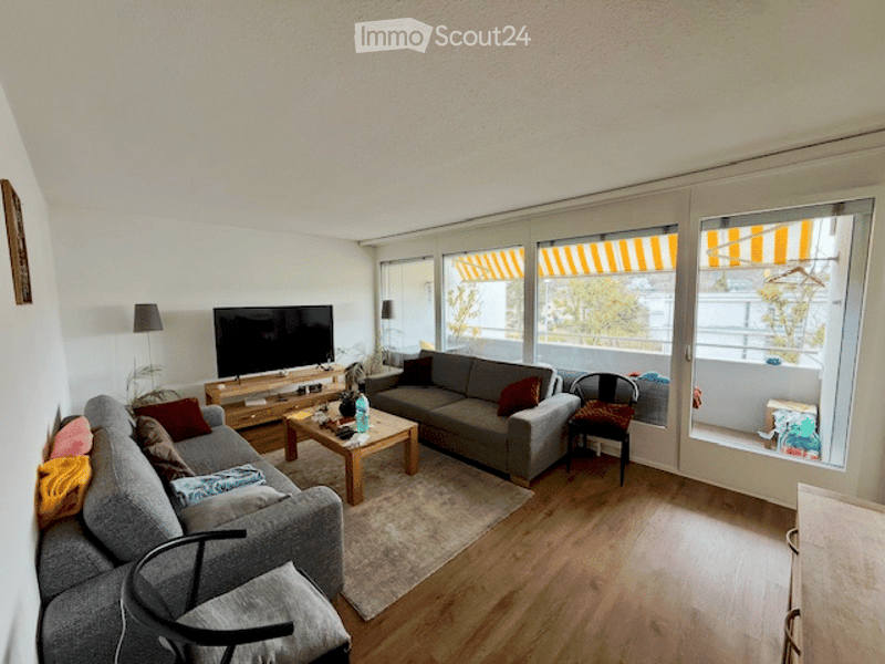 Bright living room with large windows and a sliding glass door leading to a balcony. Features a grey sectional sofa, coffee table, TV, and a small dining area with a chair.