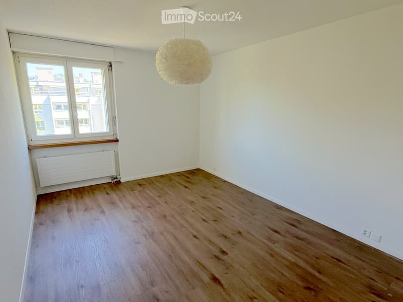 Empty room with light wooden flooring, white walls, a large window with a view, a radiator, and a hanging spherical light fixture.