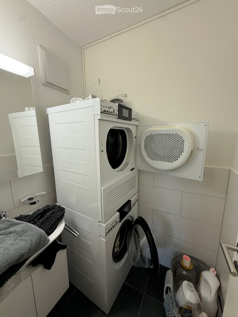 Compact bathroom with a stacked washing machine and dryer unit, towels on a shelf, and detergent bottles nearby.