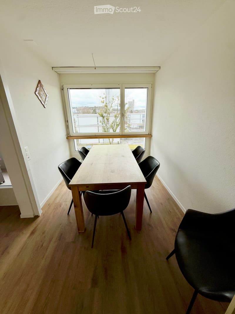A small, well-lit room with wooden flooring and white walls. A wooden table is surrounded by black chairs, positioned near a large window offering a view of the outside.