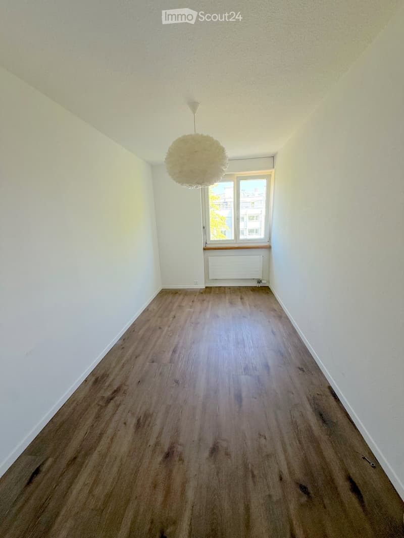 Empty hallway with wooden flooring, white walls, and a hanging spherical light fixture. A window with a radiator below it is visible at the far end.