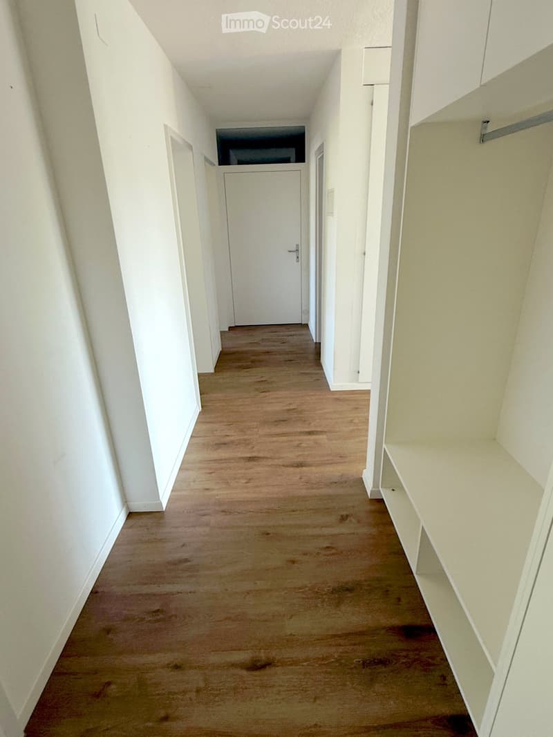 Empty hallway with light wooden flooring, white walls, and built-in shelving on one side. A closed door is visible at the end of the hallway.