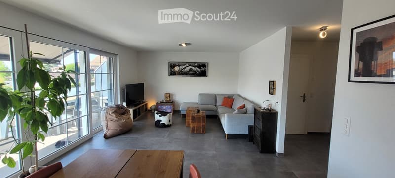 Modern living room with large windows, grey sectional sofa, coffee table, wooden side table, beige bean bag, and a television on a stand.
