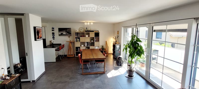 Open-plan living and dining area with large windows and a balcony. Includes a wooden dining table with chairs, a small bar area with stools, shelves with books and decor, and a potted plant near the balcony entrance.