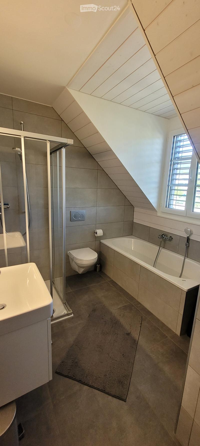 Modern bathroom with a bathtub, toilet, sink, and shower area. Features a window with blinds, tiled walls, and a dark grey bath mat on the floor.