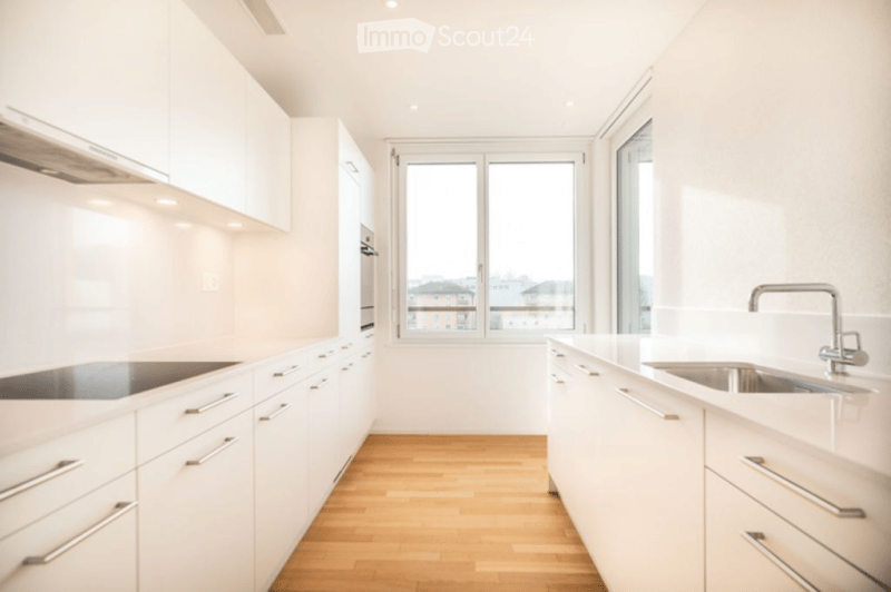 Modern kitchen with white cabinets, stainless steel appliances, wooden flooring, and large windows offering a city view.