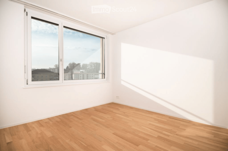 Empty room with white walls, wooden flooring, and large windows offering a view of the city.