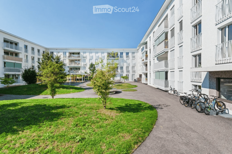 Modern multi-story residential building with balconies, surrounded by green spaces and bicycles parked along the pathway.