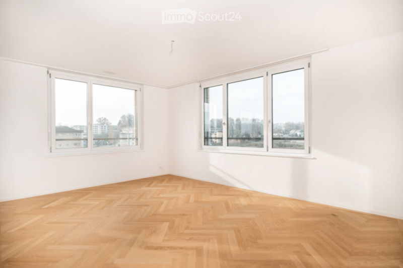 Empty room with white walls, wooden herringbone flooring, and large windows offering a view of the city and trees.