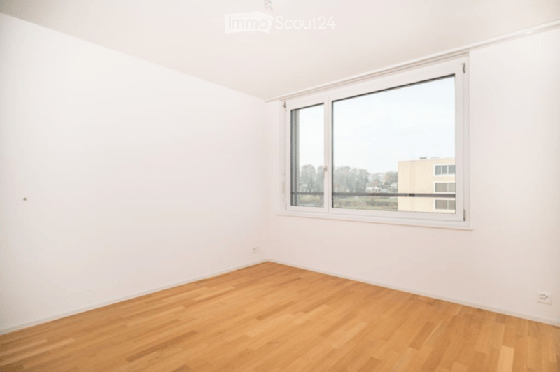 Empty room with white walls, wooden floor, and large window showing a view of buildings and trees.