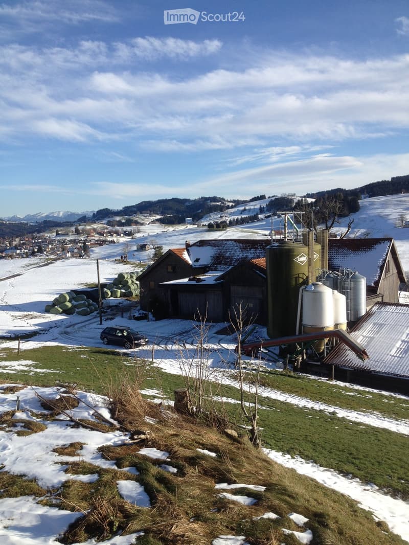 A farm complex with silos, barns, and a parked car in a snowy landscape with distant mountains.