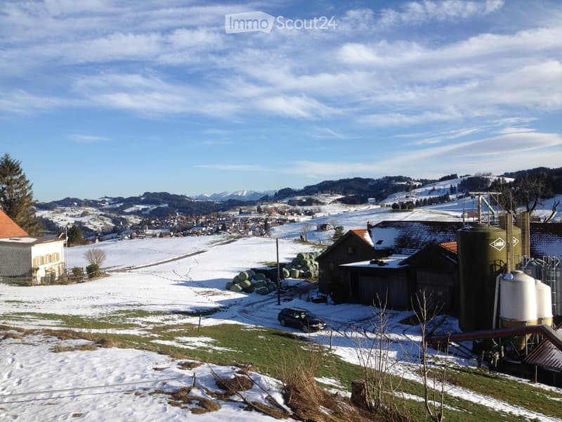 A rural landscape with snow-covered fields, farm buildings, silos, and a road leading to a village in the distance.