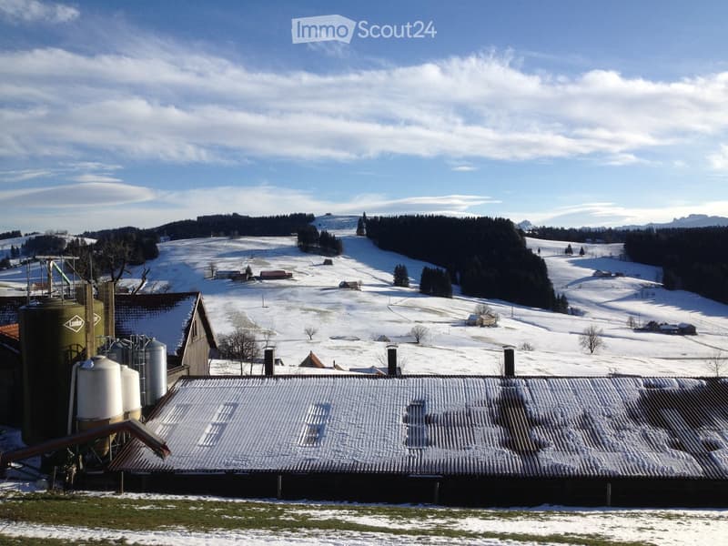 A snow-covered rural landscape with a large barn and silos in the foreground, surrounded by snow-covered fields and distant forested hills.