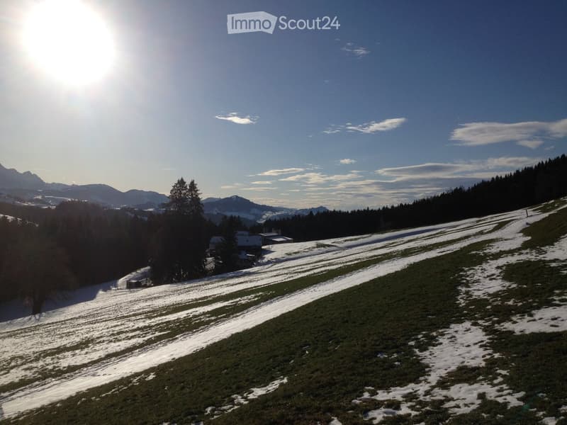 A serene, snow-dusted hillside with a small house nestled among trees, surrounded by a vast, open landscape under a clear blue sky with scattered clouds and distant mountain peaks.