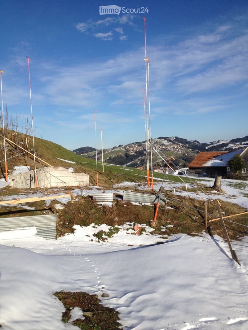 Outdoor area with snow-covered ground, wooden planks, metal structures, and distant hills and houses.
