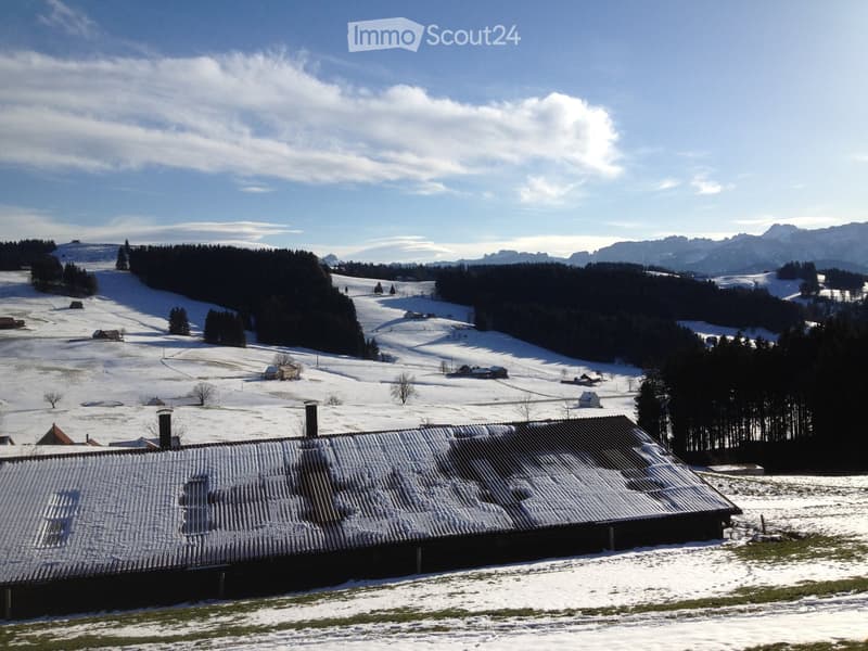 A rustic wooden building with a snow-covered roof, situated in a snowy landscape with scattered trees and distant mountains.