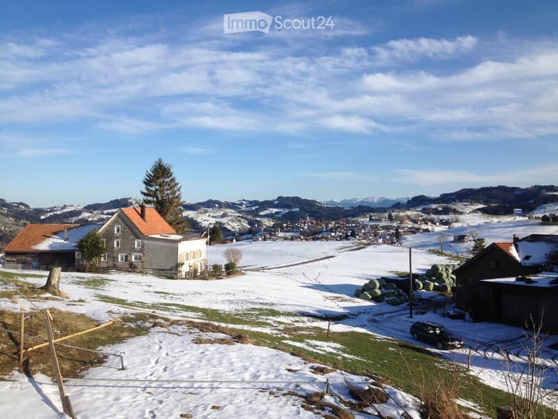 A rural house with a red-tiled roof and white walls, surrounded by snow-covered fields and distant mountains.