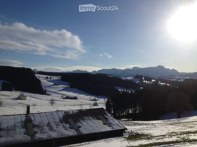 A serene winter landscape featuring snow-covered fields, scattered houses, dense forests, and distant mountain peaks under a clear blue sky.
