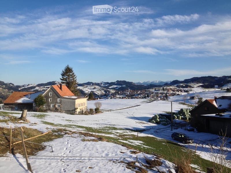 A rural village scene with several houses, snow-covered fields, and a clear blue sky. The houses have traditional architecture with pitched roofs and are surrounded by trees and open land.
