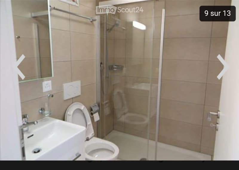 Modern bathroom with a white sink, toilet, glass-enclosed shower, and a mirror above the sink. The walls are tiled in a light brown color, and the floor is also tiled.