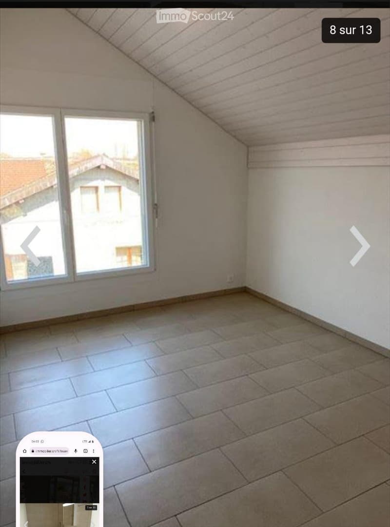 Empty attic room with exposed wooden beams, large window, and tiled floor.