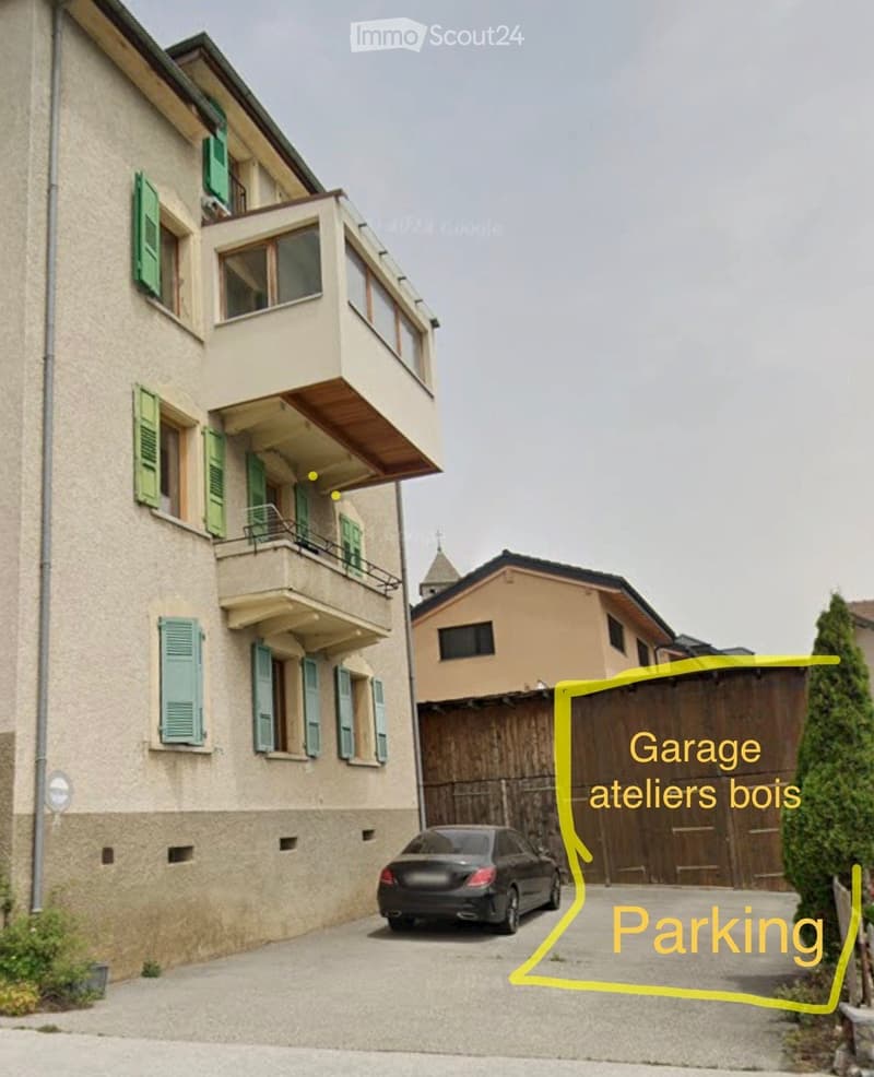 Two-story residential building with balconies and green shutters, adjacent to a garage and parking area.