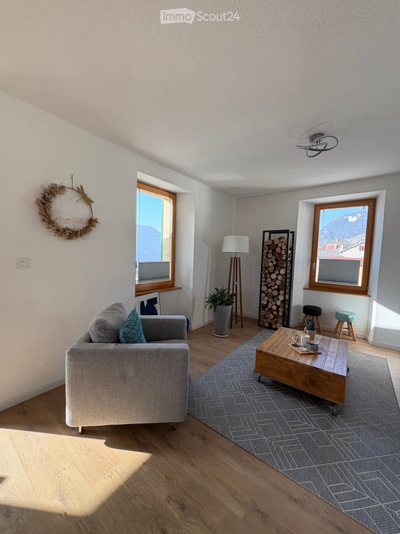 Modern living room with light wooden flooring, grey sofa, wooden coffee table, floor lamp, and two stools near a window with a scenic mountain view.