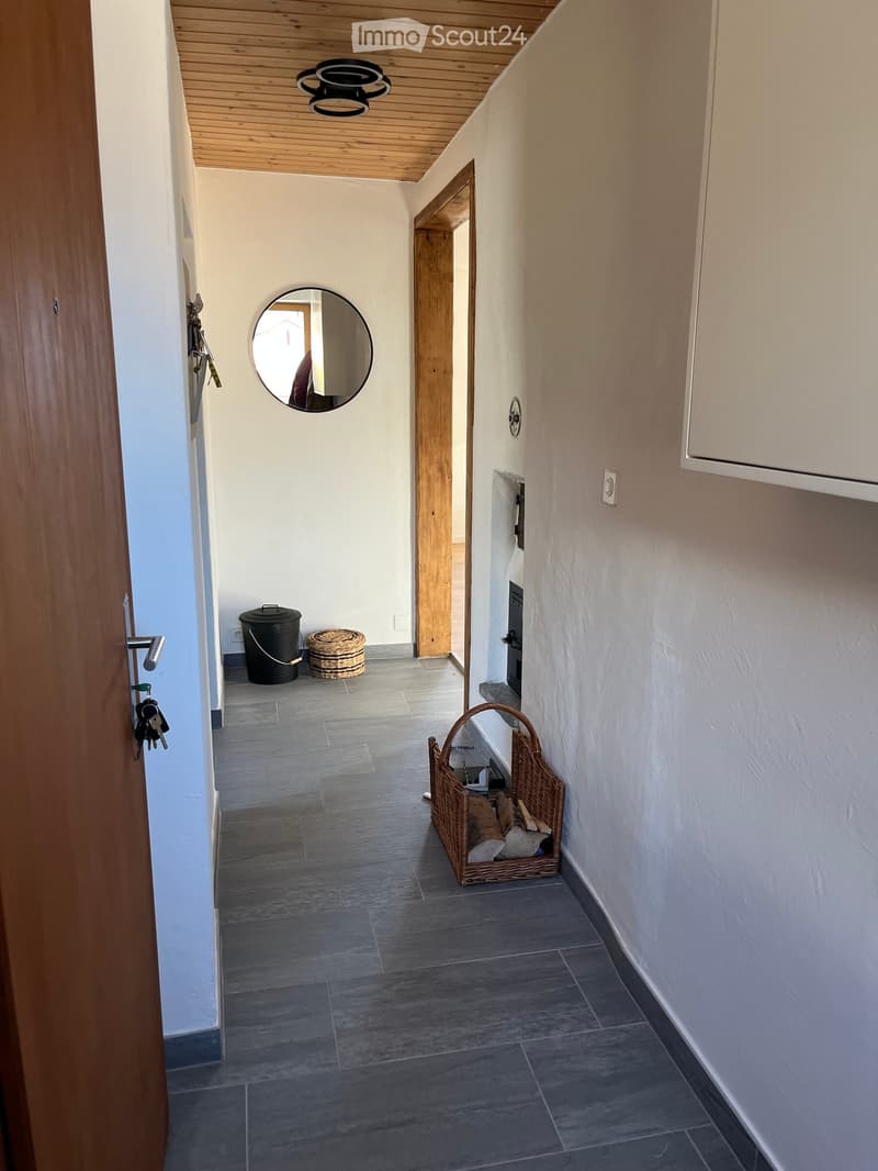 Clean hallway with wooden ceiling, grey tiled floor, and a round mirror on the wall. A wicker basket and a black trash bin are placed along the walls.