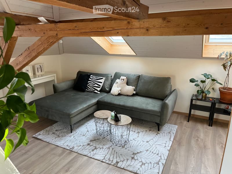 Modern living room with exposed wooden beams, grey sectional sofa, two small round tables, side table with plants, and a white rug.