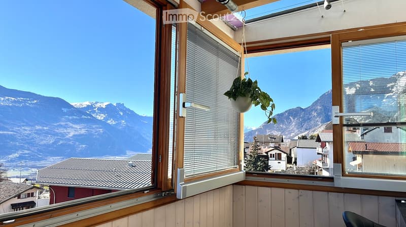 Balcony with large windows offering a scenic mountain view, featuring a hanging potted plant and wooden interior trim.
