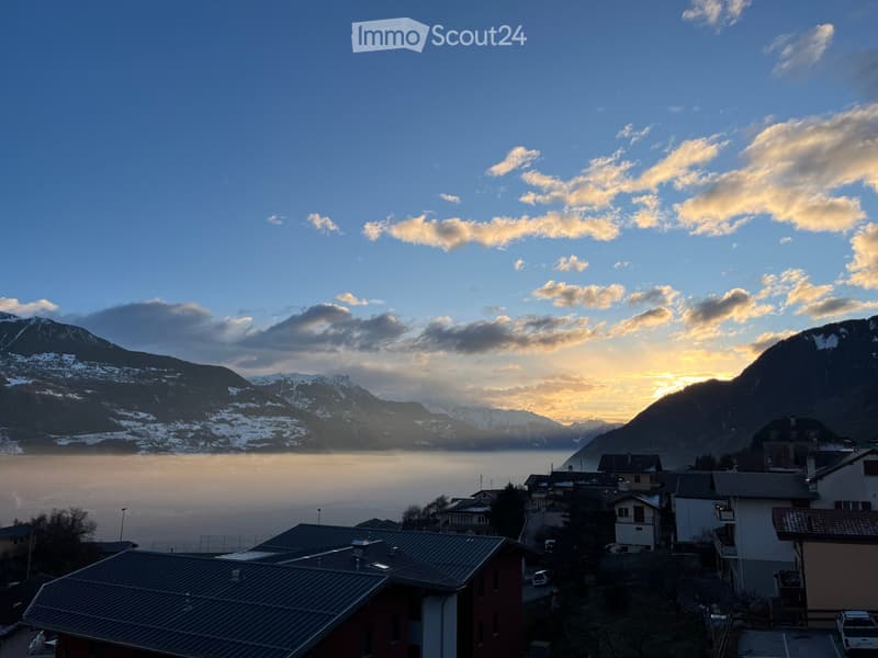 View of a village with houses and a lake surrounded by snow-capped mountains at sunset.
