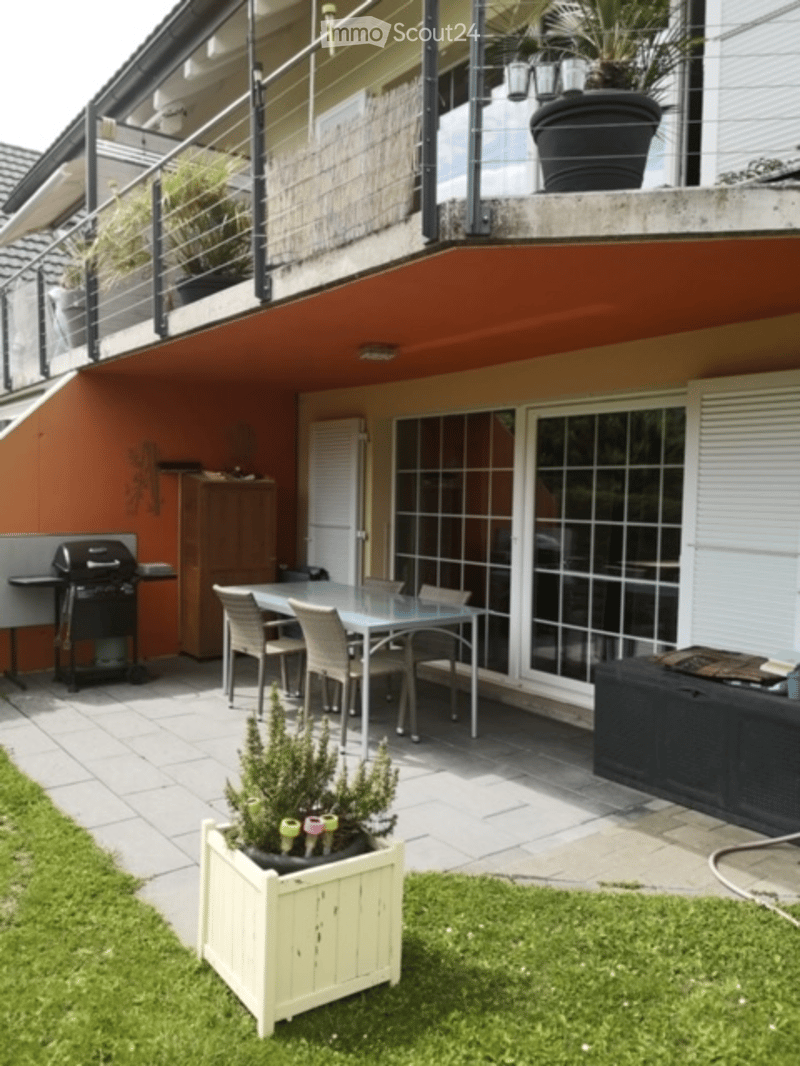 Outdoor patio area with a dining table and chairs, barbecue, potted plants, and a raised balcony above.