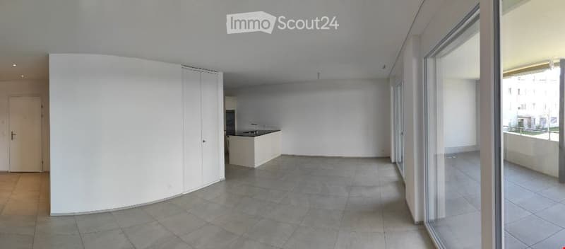 Open-plan unfurnished room with tiled flooring, white walls, large windows, and a small kitchenette area.