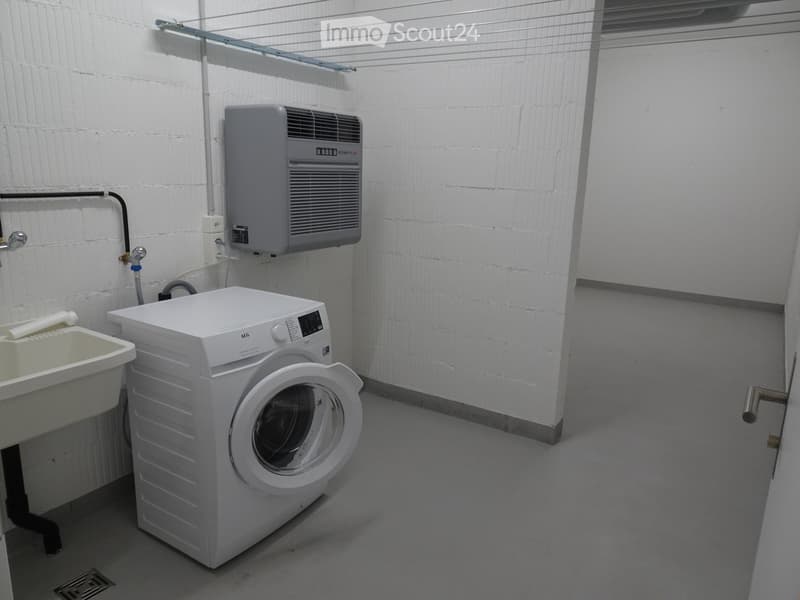 A small utility room with a white washing machine, a sink, and an air conditioner mounted on the wall. The room has white brick walls and a concrete floor.