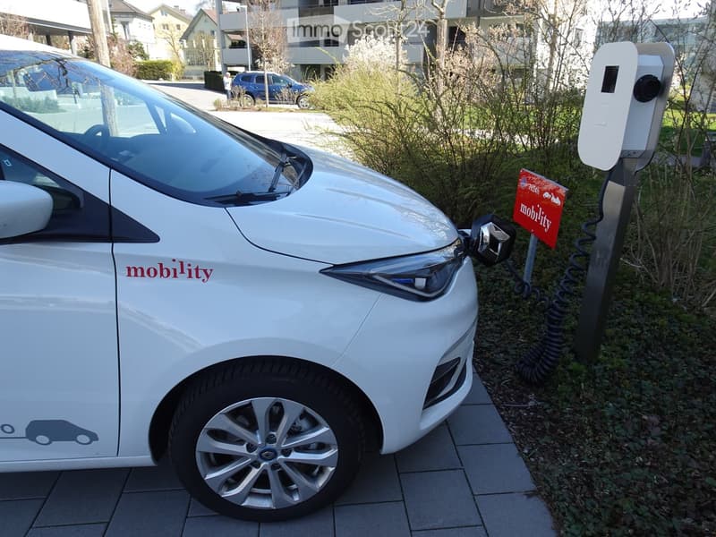 A white electric car is plugged into a charging station on a paved sidewalk, with a red sign reading "mobility" nearby.