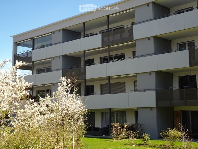Modern multi-story apartment building with balconies, large windows, and contemporary design, surrounded by greenery and flowering trees.