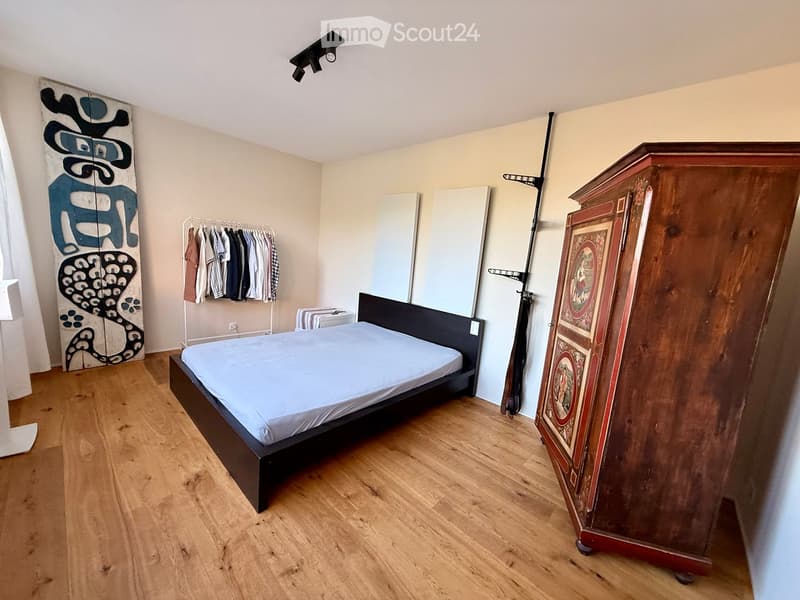 Bedroom with a single bed, a large antique wardrobe, a clothing rack, and a mirror. The room has wooden flooring and modern lighting.