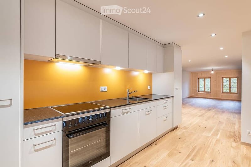 Modern kitchen with white cabinets, stainless steel appliances, and a bright orange accent wall. The adjacent room features wooden flooring and large windows.