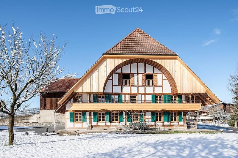 Traditional wooden house with a steep roof, green shutters, and a balcony overlooking a snowy landscape.