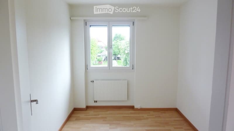 Empty room with light-colored walls, wooden flooring, a window with white frame, and a radiator below the window.