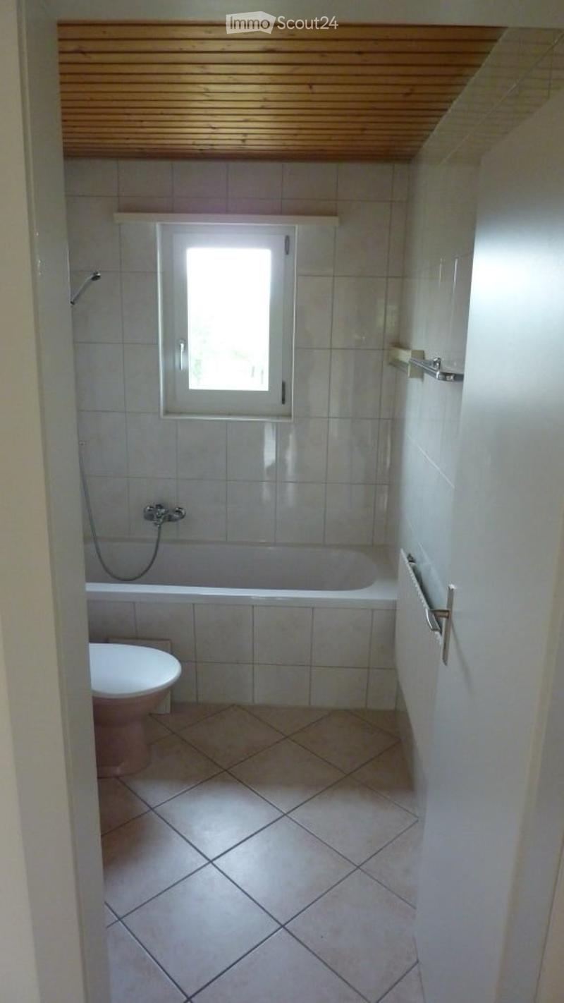 Bathroom with tiled walls and floor, a bathtub, toilet, and a small window above the bathtub. Wooden slatted ceiling.