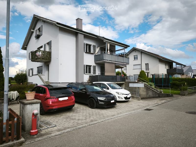 Two-story house with white exterior, multiple windows, balconies, and a small garden area. Three cars are parked in front of the house.