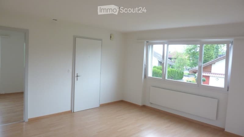 Empty room with wooden flooring, white walls, a door, and a large window with a view of greenery outside.