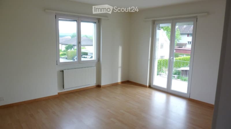 Empty room with wooden flooring, white walls, large windows, and a radiator on one wall. The room has a sliding glass door leading to a balcony or outdoor area.