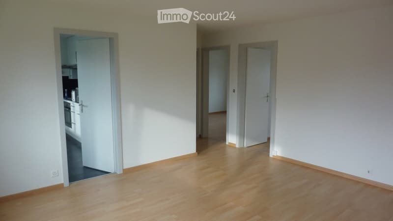 Empty room with light wooden flooring and white walls. Doors lead to other rooms, including a kitchen area visible in the background.
