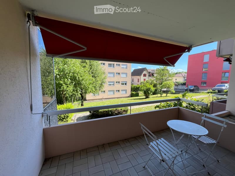 Balcony with tiled floor, white table and chairs, red awning, and view of greenery and buildings.